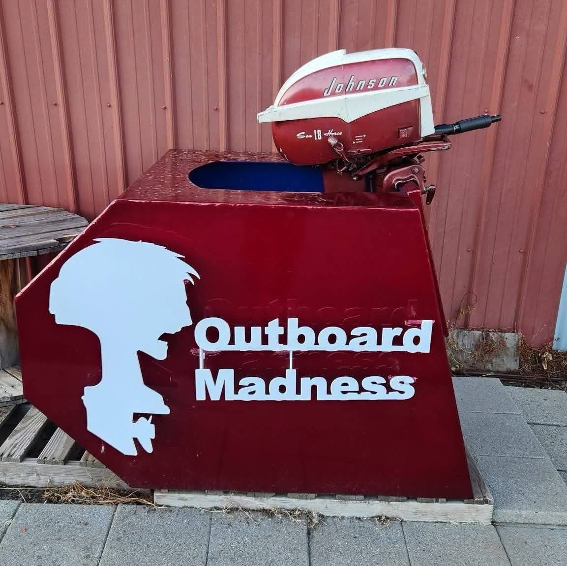 The Outboard Madness sign: a maroon stand with a white outboard-motor silhouette and the shop name in white letters, topped by a vintage red and cream outboard, in front of a red metal wall.